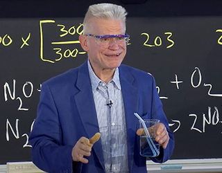 A photograph of Dr. Dolhun standing at the chalkboard, wearing safety glasses and holding a small beaker containing a pale blue solution. There are two straws in the beaker.